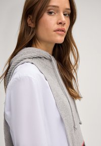 Grau gestrickter Kapuzenpullover mit strukturierter Oberfläche, ausgestattet mit einem Kordelzug und einem darunter getragenen leichten, glatten weißen Hemd. Das Modell hat lange Haare.