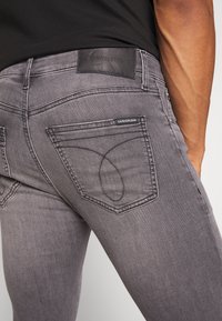 Jeans de denim gris con un corte slim. Presentan un parche de cuero con marca en la cinturilla y un stitching distintivo en los bolsillos traseros.
