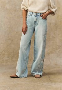 Jeans de talle ancho en azul claro con patrones florales bordados en la pierna izquierda, combinados con un suéter de punto crema y zapatos beige.