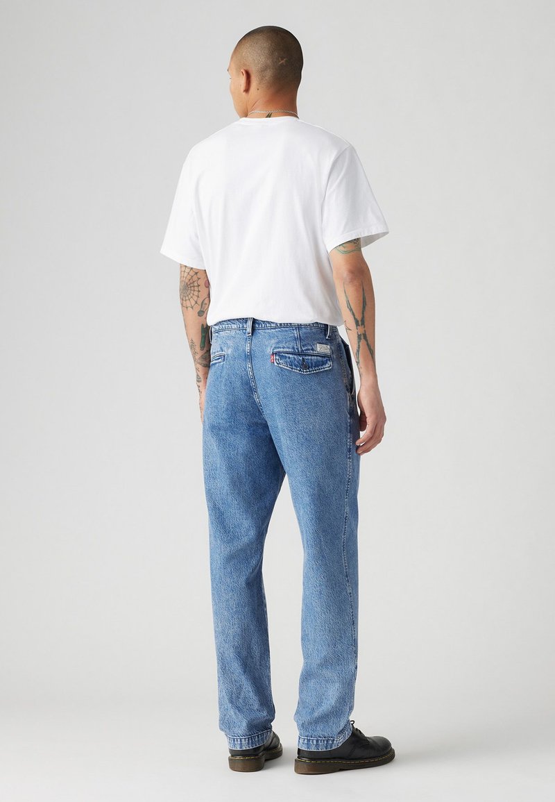 Weißes T-Shirt mit kurzen Ärmeln und blaue lässige Jeans. Die Jeans haben Taschen und eine leichte Abnutzung, während schwarze Schuhe das Outfit abrunden.