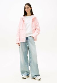 Giacca impermeabile rosa con cappuccio, felpa bianca con collo tondo sotto, jeans a gamba larga blu chiaro e scarpe bianche con accenti discreti.