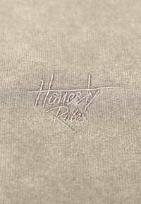 Ljusgrått textil med stickad textur, prydd med broderad "Ärlighet Regler" i en kontrasterande rosa tråd.