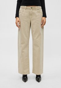 Άτομο που φοράει μπεζ παντελόνι με φαρδιά μπατζάκια, μαύρο μακρυμάνικο μπλουζάκι και μαύρα μποτάκια με μυτερή μύτη, στέκεται μπροστά σε απλό φόντο.