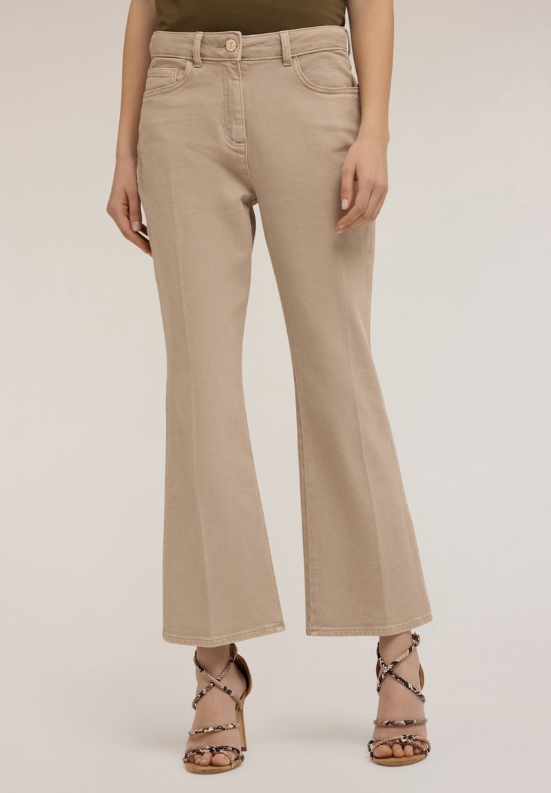 Jeans beige acampanados con una textura suave, diseño de cinco bolsillos, cierre de botón y sutil detalle de costura, combinados con sandalias de tacón alto con tiras.