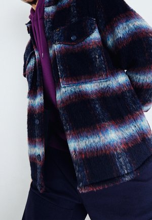 Osoba nosząca puszystą, wielokolorową kratowaną kurtkę na fioletowej bluzie z kapturem i sznurkami oraz ciemne spodnie.