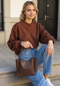 Braune Leder-Tote mit kurzen Griffen, glatter Oberfläche und offenem Design. Getragen von einer Person in einem braunen Pullover und blauen Jeans.