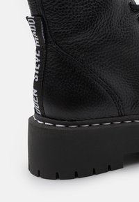 Bottines en cuir noir avec surface texturée, semelle noire épaisse et couture blanche. Présente une languette latérale avec le nom de la marque imprimé.