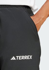 Crne TERREX hlače s glatkom teksturom, s bočnom džepom i bijelim logotipom na donjoj lijevoj nozi. Prikazan elastičan pojas.