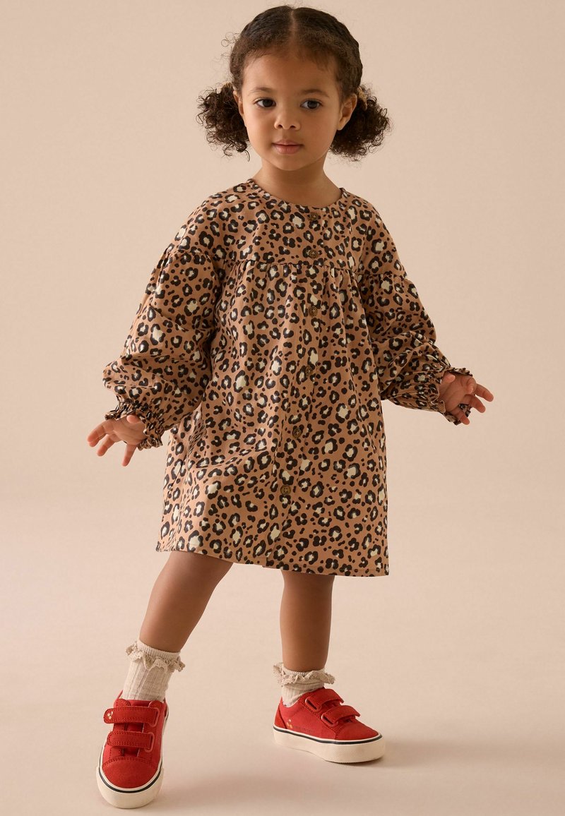 Leopardenmuster-Kleid in Beige und Braun, mit langen Puffärmeln und geraffter Taille. Kombiniert mit roten Schuhen und weißen Rüschen-Socken.