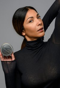 Vasca di metallo rotonda etichettata "Poudre de Riz", tenuta in una mano. L'etichetta presenta un testo in rilievo. Modella che indossa un dolcevita aderente e trasparente di colore nero.