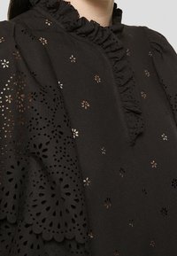 Vêtement noir avec découpes florales œillet, col montant à volants et détails en dentelle à bords festonnés superposés sur l'épaule.