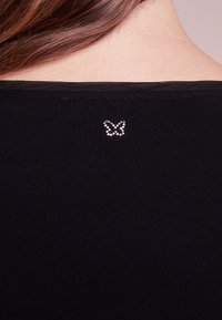 Schwarzes, geripptes Oberteil mit einem kleinen, mit Kristallen besetzten Schmetterlingsdesign am Rückenausschnitt.