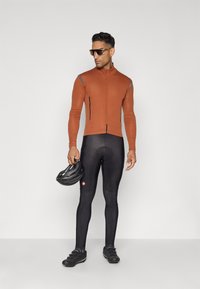 Tenue de cyclisme comprenant un maillot à manches longues ajusté orange et un cuissard noir avec des accents réfléchissants. Le mannequin tient un casque noir.