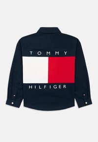 Μπλε ναυτικό πουκάμισο με γιακά, με μεγάλο λευκό και κόκκινο μπλοκ σχέδιο στην πλάτη και το κείμενο «TOMMY HILFIGER» σε έντονα λευκά γράμματα.