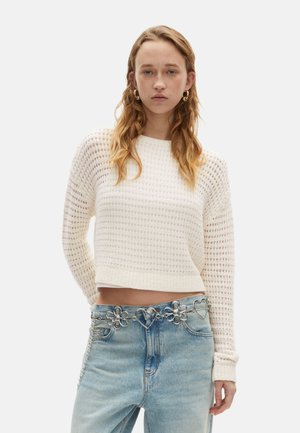 Witte cropped trui met een gestructureerde weving en lange mouwen, gecombineerd met lichtblauwe jeans voorzien van metalen bloemen- en hartversieringen.
