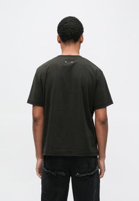 Σκούρο πράσινο βαμβακερό μπλουζάκι με χαλαρή εφαρμογή, κοντά μανίκια, στρογγυλεμένο λαιμό και δύο μεταλλικά αεροπλάνα κοντά στον πίσω λαιμό.