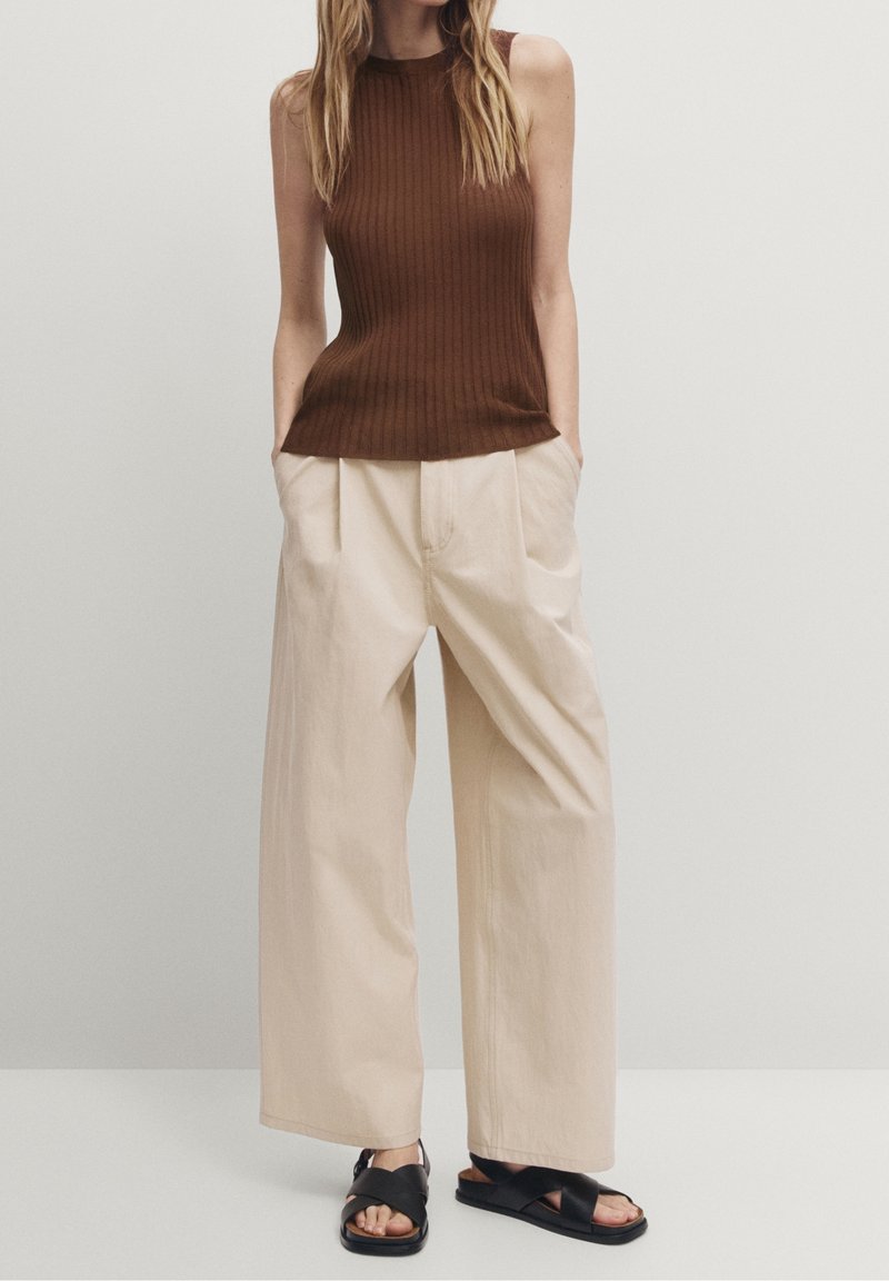 Ruskea ribbiprintillinen hihaton paita, yhdistettynä beigen värisiin leveälahkeisiin housuihin; asun täydentävät mustat sandaalit. Yksinkertainen malli rennolla istuvuudella.