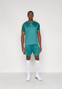 Sporta komplekts, kas sastāv no tirkīzzila krekla ar melnām pleciem un neona zaļām detaļām, saskaņotām šortiem, baltām zeķēm un sporta apaviem, viss paredzēts sportam.