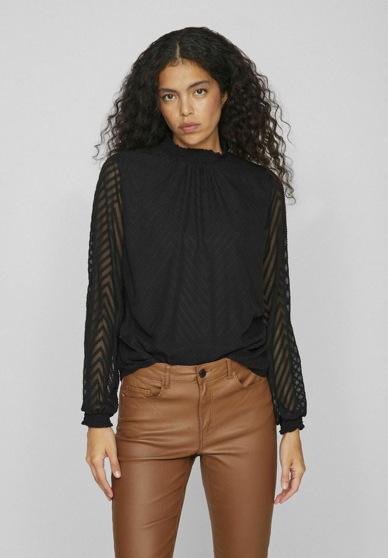 Blusa nera a maniche lunghe con maniche trasparenti strutturate e colletto alto, abbinata a pantaloni in pelle marrone aderenti. Tessuto liscio con motivo a zigzag.