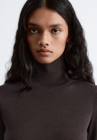 Brown turtleneck sweater with a smooth, soft texture and subtle ribbed details along the neckline and cuffs. Close-up of the design.