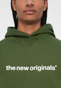 Olive zelena kapuca iz bombaža s kengurujskim žepom. Na sprednji strani se nahaja beli napis "the new originals". Ribsani detajli na kapuci.