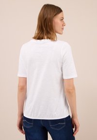 Kvindelig med skulderlangt lysebrunt hår iført en ensfarvet hvid t-shirt med korte ærmer og mørkeblå jeans, stående siden til mod en beige baggrund.