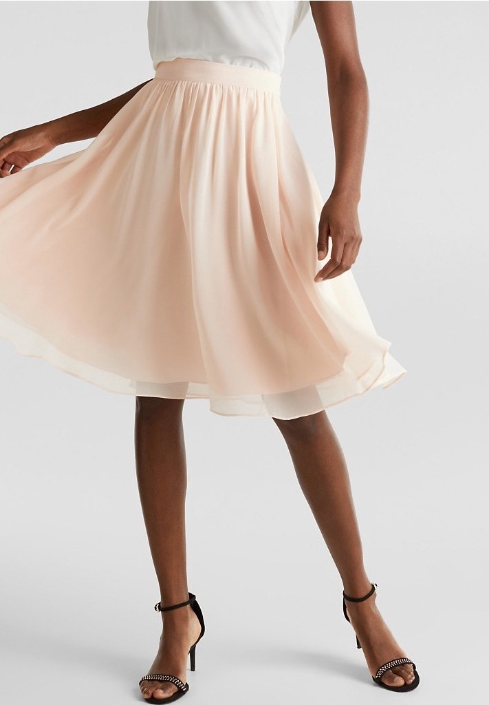 Femme portant une jupe midi fluide rose clair et des sandales à talons noirs, debout avec une main soulevant doucement la jupe.