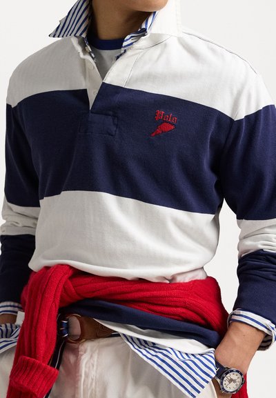 Marineblå og hvidstribet rugbytrøje med et rødt broderet logo, lagdelt over en blåstribet skjorte med krave, parret med en rød sweater knyttet om taljen.