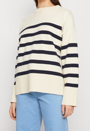 Femme portant un pull crème à rayures horizontales bleu marine et un pantalon en velours côtelé bleu clair, visible des épaules jusqu'aux hanches.