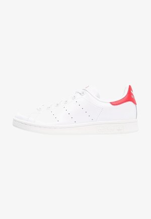 Biały niskoprofilowy sneaker Adidas Stan Smith z czerwonym paskiem na pięcie, perforowanymi bocznymi paskami i białą gumową podeszwą.