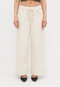 Pantalones de pierna ancha beige con cinturilla elástica y cordón. Incluye bolsillos laterales y tiene una textura suave y lisa.