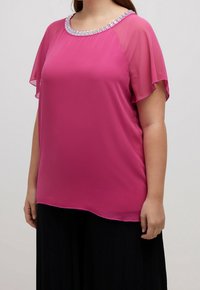 Blouse magenta avec manches transparentes et encolure ornée de bijoux, dotée d'une coupe ample et d'une texture lisse, portée sur une jupe plissée foncée.