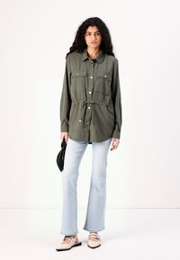 Olijf groene button-up shirt met twee borstzakken, strik taille en lange mouwen. Gecombineerd met lichtblauwe flare jeans en beige instappers.