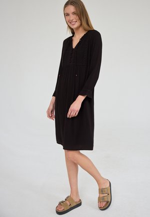 Młoda kobieta uśmiecha się, ubrana w czarną, długorękawową sukienkę do kolan z haftem oraz brązowe sandały na klamry, stoi na neutralnym tle.