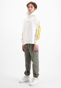 Witte hoodie met geel vlamontwerp op de mouwen, gecombineerd met groene cargobroek met zijzakken en witte sneakers.