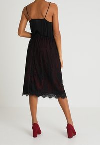 Bodice in satin nero con spalline sottili, che si trasforma in una gonna in pizzo bordeaux plissettata. Orlo scallopato notevole e tacchi a blocco rossi.