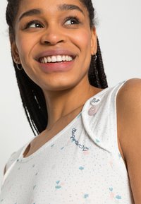 T-shirt branca sem mangas com padrões florais em azul e rosa. Apresenta botões decorativos e texto em caligrafia no ombro. Material de algodão suave.