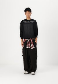 Black sweatshirt with white graphic text, oversized black pants, and a red plaid flannel tied around the waist. Grey sneakers complete the look.