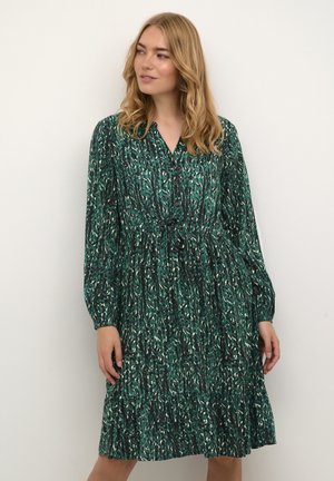 Frau mit blonden Haaren, die ein langes, grün gemustertes Hemdblusenkleid trägt, stehend und zur Seite schauend vor einem schlichten Hintergrund.
