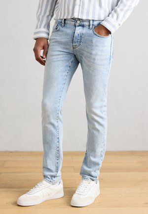 Mand iført lyseblå slim-fit jeans, hvide sneakers og en blå- og hvidstribet langærmet skjorte, stående på trægulv.