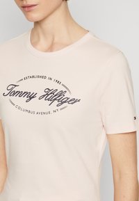 Světle růžové bavlněné tričko s kulatým výstřihem, krátkými rukávy a tmavě modrým logem "Tommy Hilfiger". Malý detail loga na rukávu.