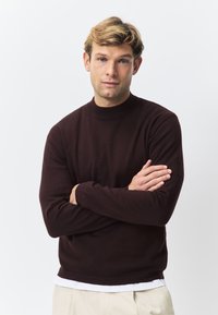 Bourgogne strikket sweater med høj hals, lange ærmer og ribbet manchetter. Glat tekstur, figursyet design, brugt over en hvid skjorte.