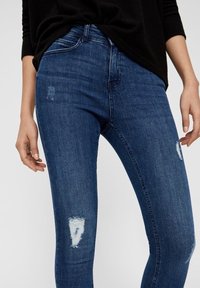 Jeans de mezclilla azul ajustados con un diseño desgastado. Presentan pequeños desgarros en las rodillas, forma ceñida al cuerpo y un estilo clásico de cinco bolsillos.