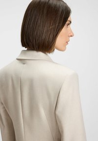 Beige bleiseri, jolla on sileä rakenne, lovellinen kaulus ja tuuletus yksityiskohtia. Ominaisuutena minimalistinen muotoilu ja hienovaraiset metalliset yksityiskohdat takana.