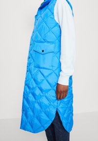 Gewatteerd blauw mouwloos gewatteerd vest met drukknoopsluiting en een voorzak, voorzien van een hoge kraag en zijopeningen voor bewegingsvrijheid.