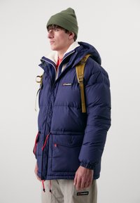 Doudoune bleu marine avec un col en peluche blanche, cordons de serrage rouges ajustables et poches avant. Portée avec un bonnet vert et un sac à dos beige.