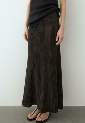 Frau in einem langen, dunklen plissierten Rock, schwarzem Oberteil und schwarzen Zehensandalen, steht vor einem schlichten hellen Hintergrund.