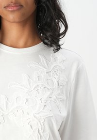 Witte katoenen t-shirt met complexe witte bloemenborduurwerk aan de linkerkant, voorzien van een ronde halslijn en korte mouwen.