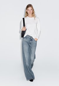 Weißes Langarmhemd, hellblaue weite Jeans und schwarze Tasche. Stiefel mit spitzem Zeh. Glattes Material mit minimalen Details.