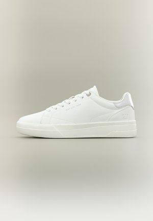 Biela nízka teniska so šnúrkami, nenápadným označením na boku a päte a gumovou podrážkou na neutrálnom béžovom pozadí.